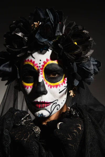 Retrato de mujer en guirnalda y maquillaje de catrina mirando a la cámara sobre fondo negro - foto de stock