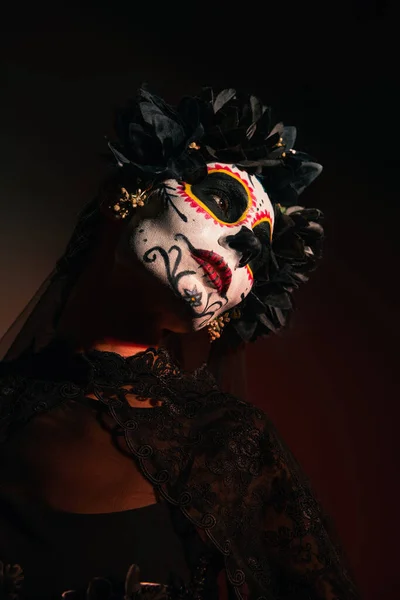 Mujer en guirnalda con flores negras y maquillaje catrina mirando hacia otro lado sobre fondo borgoña - foto de stock