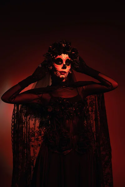 Donna in costume nero e trucco Santa Muerte toccando ghirlanda nera su sfondo bordeaux con illuminazione rossa — Foto stock