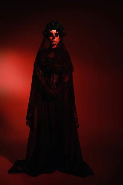 Full length of woman in mexican santa muerte costume looking at camera on burgundy background with red lighting — Stock Photo