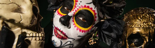 Portrait of woman in santa muerte makeup and black wreath looking at camera near skulls isolated on black, banner — Stock Photo