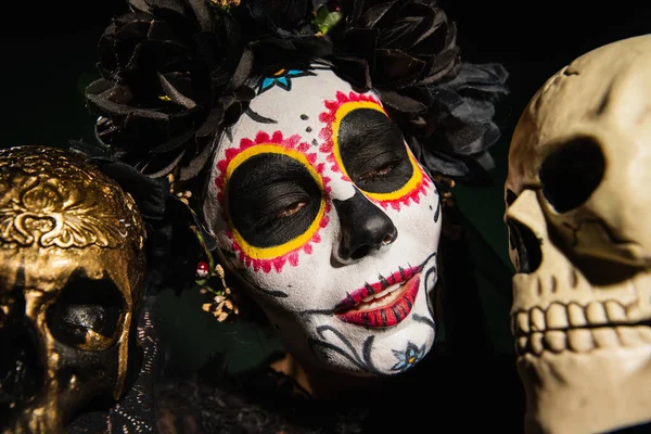 Portrait of woman in mexican santa muerte makeup looking at skull isolated on black — Stock Photo