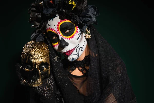 Mujer en maquillaje catrina mexicana y corona negra sosteniendo cráneo sobre fondo verde oscuro - foto de stock