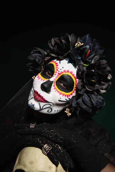 Женщина в мексиканском костюме Санта-Муэрте смотрит в камеру рядом с черепом, изолированным на черном — стоковое фото