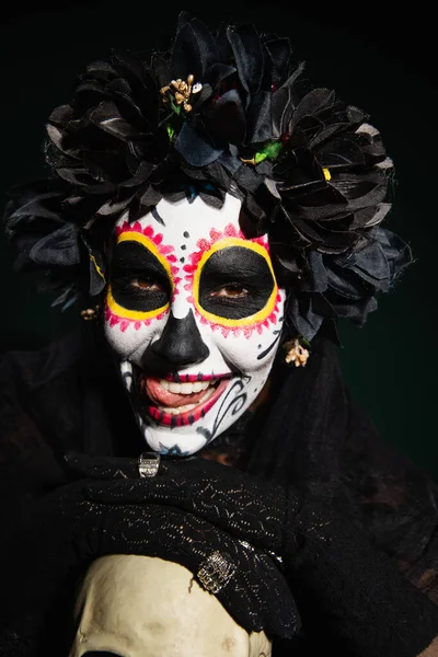 Woman in creepy day of dead costume sticking out tongue near skull on dark green background — Stock Photo