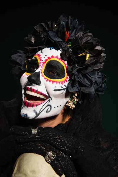 Cheerful woman with santa muerte makeup touching skull on dark green background — Stock Photo