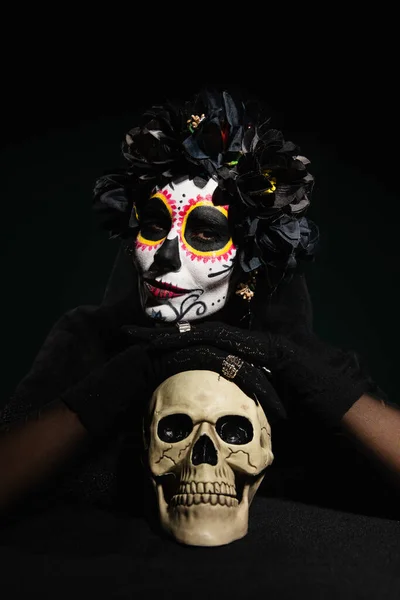 Femme en costume mexicain de jour de la mort regardant la caméra près du crâne isolé sur noir — Photo de stock