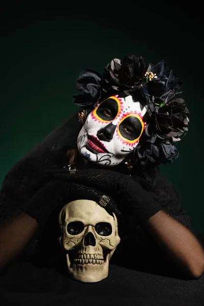 Woman in black wreath and day of death costume looking at camera near skull on dark green background — Stock Photo