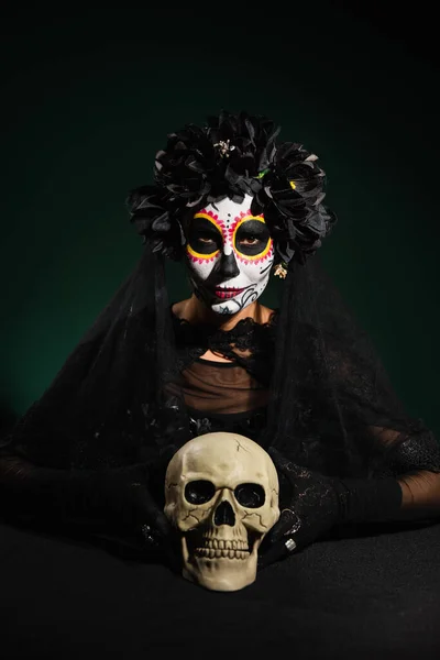 Mujer en guirnalda con velo y maquillaje catrina mirando a la cámara cerca del cráneo sobre fondo verde oscuro - foto de stock