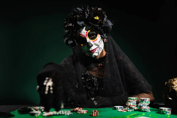 Mujer en el día de la muerte disfraz de Halloween mirando a la cámara cerca del cráneo y jugando fichas sobre fondo verde oscuro - foto de stock
