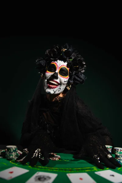 KYIV, UKRAINE - SEPTEMBER 12, 2022: Smiling woman in santa muerte costume near playing chips and cards on dark green — Stock Photo