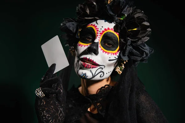 Portrait of woman in santa muerte costume and makeup holding card isolated on dark green — Stock Photo