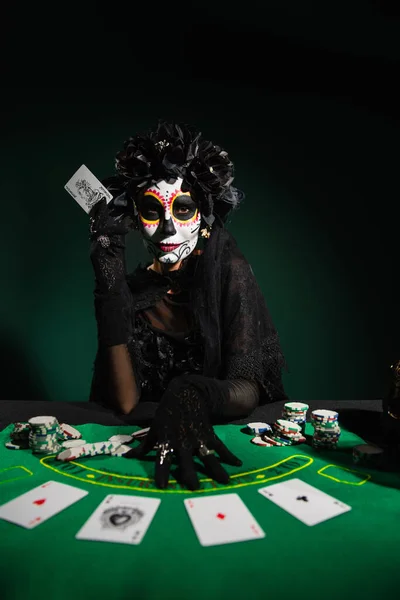 KYIV, UKRAINE - SEPTEMBER 12, 2022: Woman with sugar skull makeup and costume holding playing card on dark green — Stock Photo