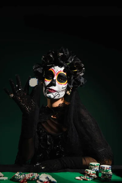 Mujer en traje de santa muerte y corona sosteniendo chip jugando sobre fondo verde oscuro - foto de stock