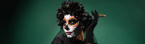 Woman with santa muerte makeup and wreath holding cigar on green background, banner — Stock Photo
