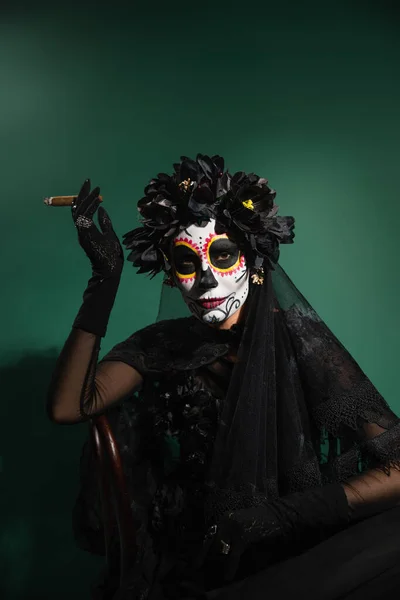 Frau im mexikanischen Santa-Muerte-Kostüm und Make-up hält Zigarre in der Hand und blickt in die Kamera auf grünem Hintergrund — Stockfoto