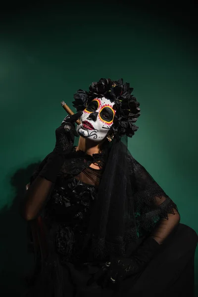 Frau in Totensonntag-Halloween-Make-up und schwarzem Kostüm mit Zigarre auf grünem Hintergrund — Stockfoto
