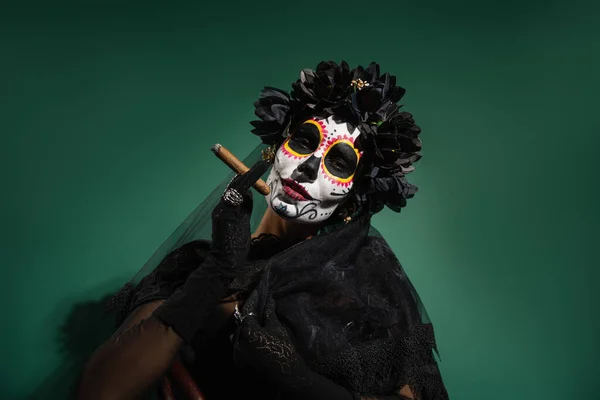 Woman in spooky halloween costume and makeup holding cigar on green background — Stock Photo