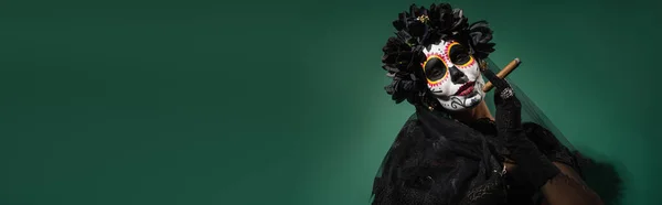 Woman in day of death halloween costume with black flowers on wreath holding cigar on green background, banner — Stock Photo