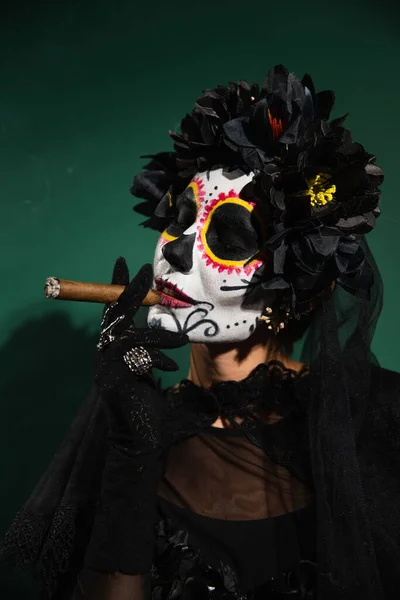 Женщина в черном венке и сахарный череп макияж курение сигары на зеленом фоне — стоковое фото