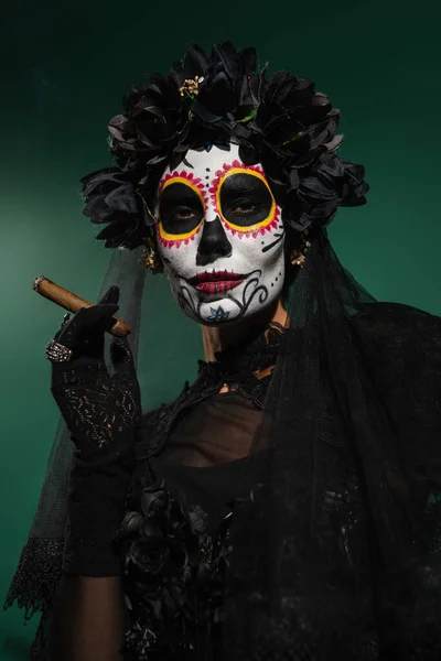 Frau in mexikanischem Day of Death Make-up und schwarzem Kostüm mit Zigarre auf grünem Hintergrund — Stockfoto