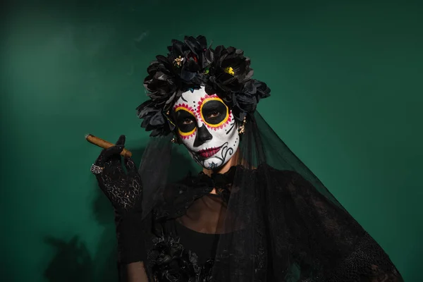 Femme dans le jour effrayant de la mort costume d'Halloween regardant la caméra et tenant cigare sur fond vert — Photo de stock