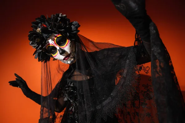 Femme en costume gothique et maquillage du crâne de sucre regardant la caméra sur fond rouge — Photo de stock