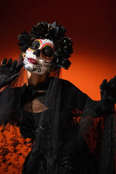 Mulher com açúcar assustador crânio maquiagem e traje preto em pé no vermelho — Fotografia de Stock
