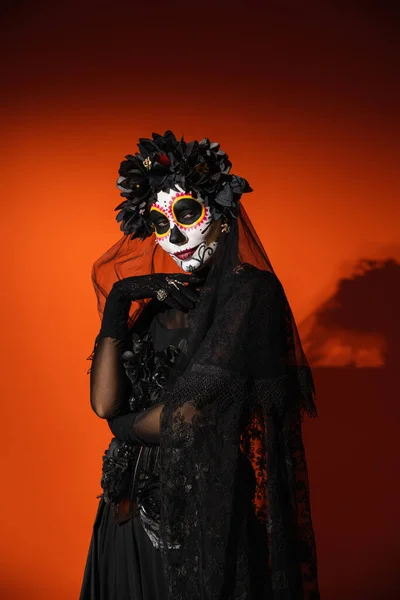 Mujer en espeluznante día mexicano de la muerte traje y maquillaje mirando a la cámara sobre fondo rojo - foto de stock