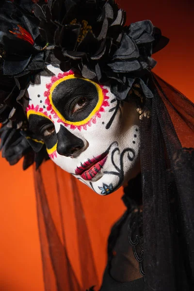 Портрет женщины в традиционный мексиканский день мертвого макияжа и черного венка, смотрящей в камеру на оранжевом фоне — стоковое фото