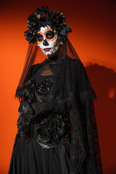 Mujer en el maquillaje de Halloween y traje con flores negras y velo de encaje sobre fondo naranja con sombra oscura - foto de stock