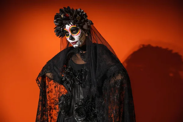 Mujer en día mexicano de maquillaje muerto y traje con velo de encaje negro posando sobre fondo naranja con sombra oscura - foto de stock