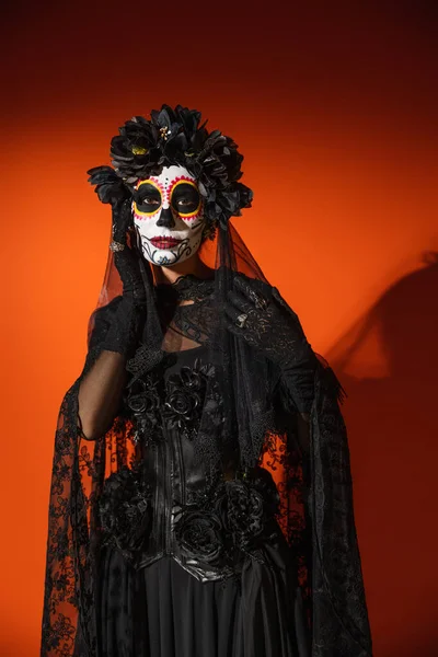 Frau in schwarzem Halloween-Kostüm und Zuckerschädel-Make-up posiert auf orangefarbenem Hintergrund mit dunklem Schatten — Stockfoto