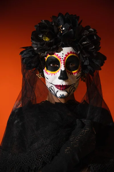 Frau in traditionellem santa muerte Make-up und schwarzem Kranz mit Schleier blickt in die Kamera auf orangefarbenem Hintergrund — Stockfoto