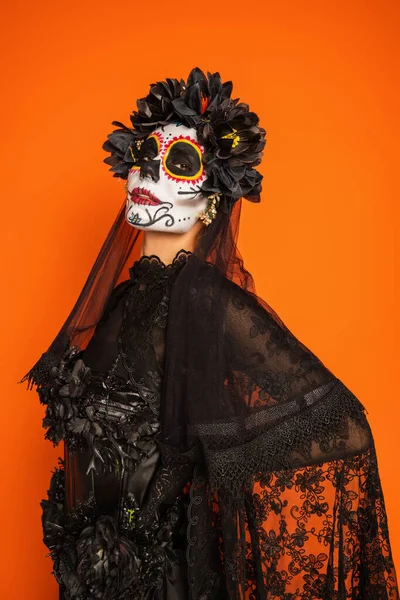 Frau in Catrina-Make-up und schwarzem Kostüm mit Kranz und Schleier blickt isoliert in die Kamera auf orange — Stockfoto