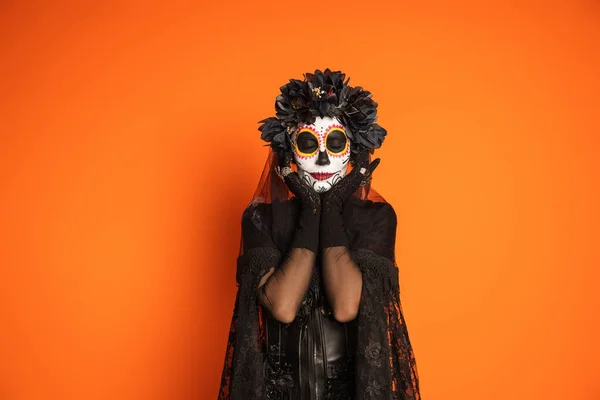 Mulher com maquiagem assustadora halloween vestindo traje preto e coroa de flores de pé com olhos fechados isolados em laranja — Fotografia de Stock