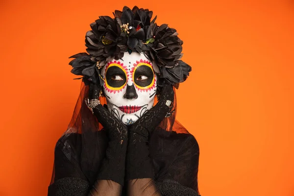 Mujer en el tradicional día mexicano de maquillaje muerto y corona negra cogida de la mano cerca de la cara y mirando hacia otro lado aislado en naranja - foto de stock