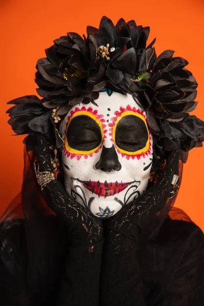 Retrato de mujer sonriente con los ojos cerrados y maquillaje de Halloween espeluznante cogido de la mano cerca de la cara aislado en naranja - foto de stock