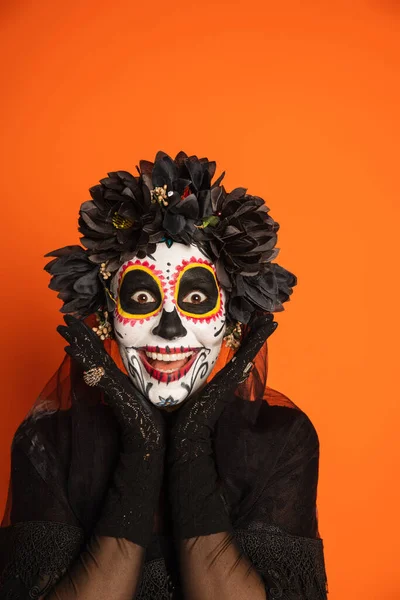 Mujer excitada en espeluznante maquillaje de halloween y corona negra cogidas de la mano cerca de la cara y mirando a la cámara aislada en naranja - foto de stock