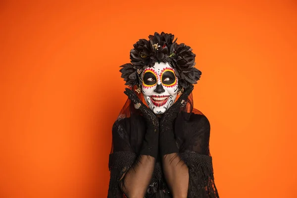 Mujer sonriente en maquillaje de cráneo de azúcar y traje de halloween negro cogido de la mano cerca de la cara aislado en naranja - foto de stock