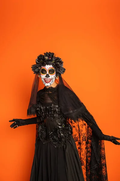 Sonriente mujer en negro bruja vestido y espeluznante halloween maquillaje aislado en naranja - foto de stock