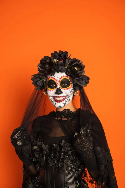 Happy woman in traditional day of dead makeup touching black veil and looking at camera isolated on orange — Stock Photo