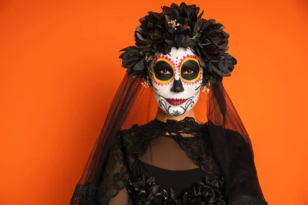 Жінка в чорному костюмі відьми і макіяжі цукрового черепа дивиться на камеру ізольовано на помаранчевому — стокове фото