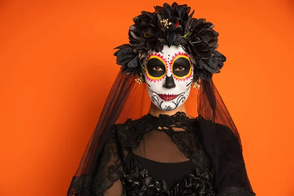 Femme en costume de sorcière noire et maquillage du crâne de sucre regardant la caméra isolée sur orange — Photo de stock