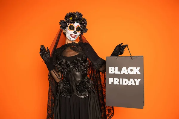 Mujer feliz en el maquillaje tradicional de santa muerte y traje de celebración de viernes negro bolsa de compras aislada en naranja - foto de stock