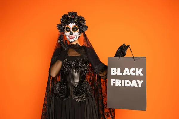 Femme heureuse dans le jour mexicain de maquillage mort et costume tenant sac à provisions noir vendredi isolé sur orange — Photo de stock