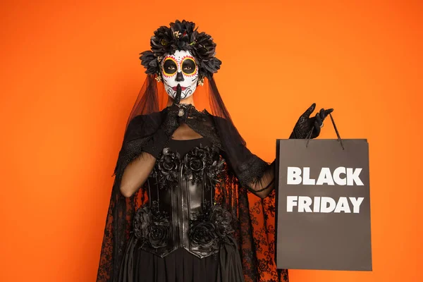 Femme dans le crâne de sucre Halloween maquillage et costume montrant le silence et tenant sac à provisions noir vendredi isolé sur orange — Photo de stock