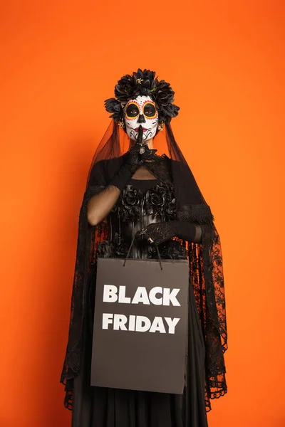 Mujer en el maquillaje espeluznante y traje de bruja sosteniendo el viernes negro bolsa de compras y mostrando el signo de silencio aislado en naranja - foto de stock