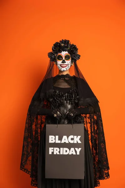 Mujer sonriente en el maquillaje de Halloween de miedo y traje de celebración de viernes negro bolso de compras aislado en naranja - foto de stock