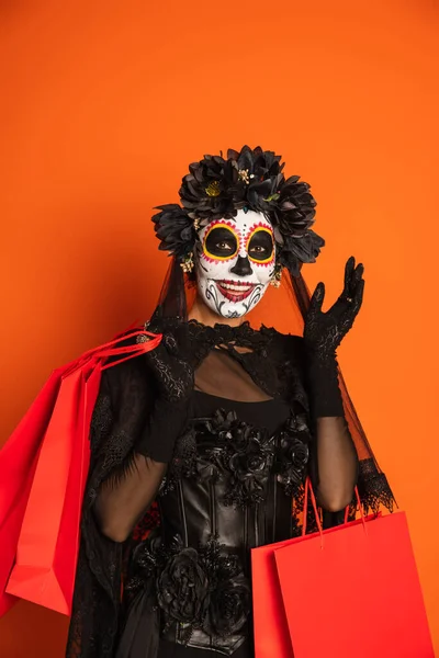 Жінка в макіяжі цукрового черепа і чорний костюм Хеллоуїна тримає сумки для покупок і махає рукою ізольовано на помаранчевому — стокове фото